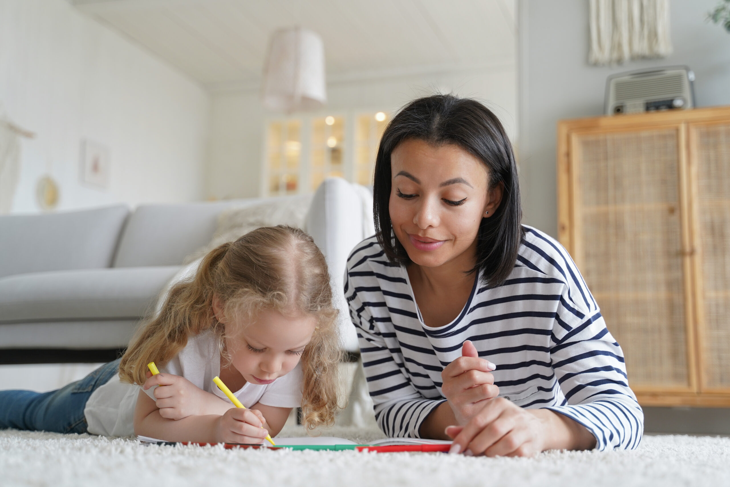 Mother and adopted daughter paint by colorful pencils, lying on floor at home. Creativity, adoption